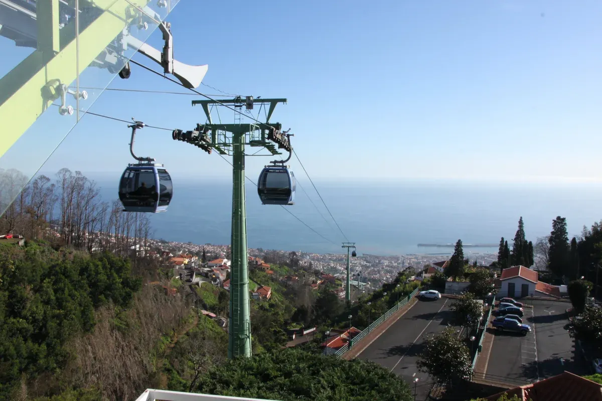 Two Funchal cable car cabins passing a support tower above the rooftops and coastline near Monte, Madeira