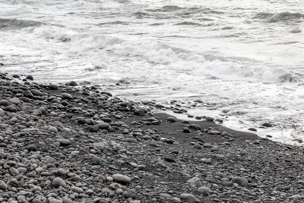 Rolled grey pebbles and a patch of black sand at the waterline of Praia Formosa in Funchal, Portugal