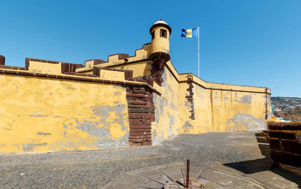 The yellow ochre battlements and corner watchtower of the Sao Tiago fort against a clear sky in Funchal, Madeira, Portugal